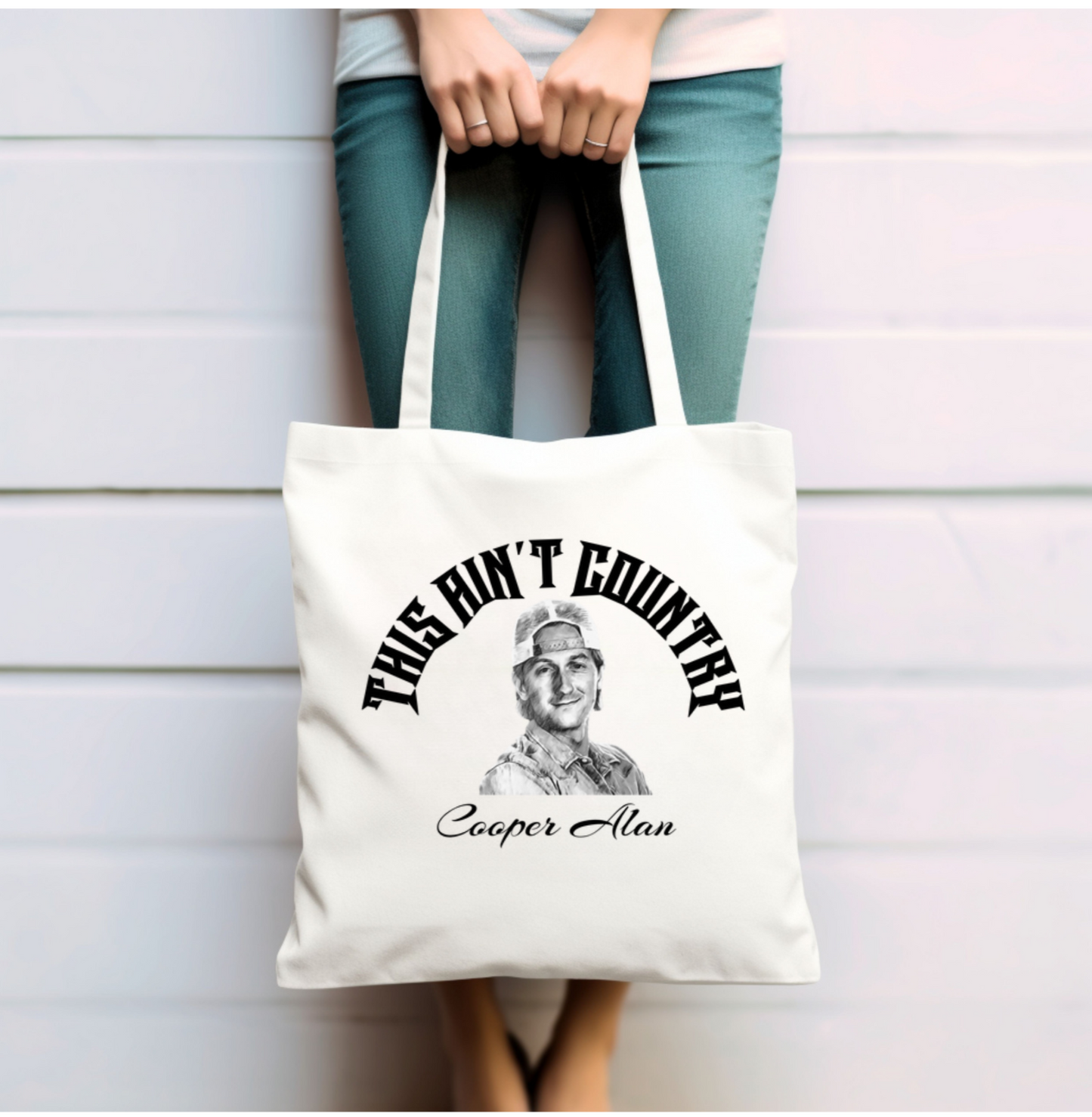 Lady hold the tote bag with wording in arch saying "This ain't country", below is a sketch drawing of Cooper Alan and below that is his name in black text