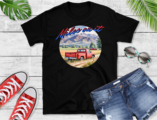 Featuring an idyllic countryside scene with majestic mountains in the backdrop and a classic red pickup truck ready to hit the road with bold “Miles on it” text arches over this serene landscape.