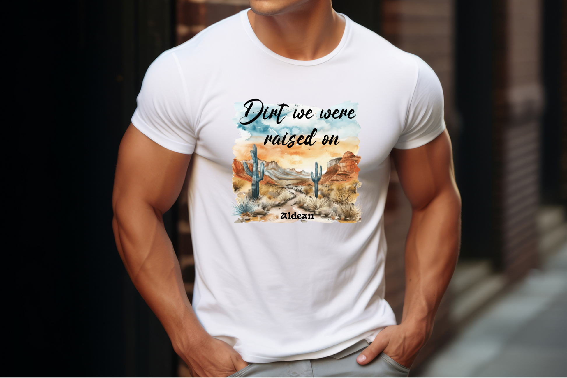 Featuring a beautifully illustrated landscape with iconic cacti and a rustic mountains. The watercolor sky adds to the image.  The phrase “Dirt we were raised on” in a casual script font at the top, and “Aldean” in a simple serif font at the bottom.