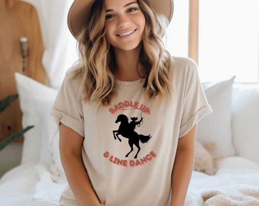 Featuring a dynamic silhouette of a cowboy riding a bucking horse, with a lasso twirling overhead. The phrase “SADDLE UP” arches above the rider in bold, western-style lettering, while “LINE DANCE” curves below. Set against a plain background, the striking black silhouette and rose-hued text.