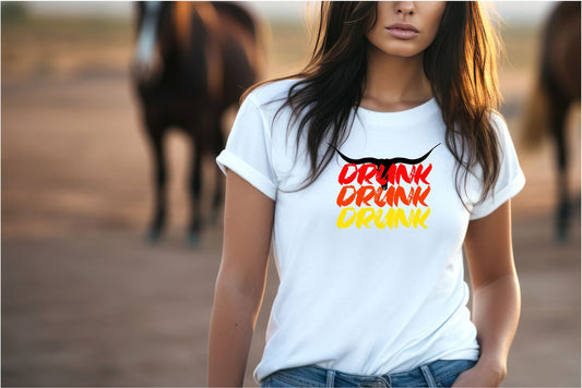 Lady standing in a rural setting wearing a white t-shirt displaying the words "Drunk Drunk Drunk" in red, orange and yellow. On top of wording is a black longhorn.