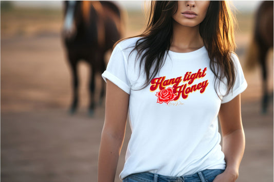 Lady standing in rural setting wearing a white t-shirt with the wording "Hang tight honey" in Red, Orange and yellow text. There is also a rose in the same colouring under the hang and next to the honey