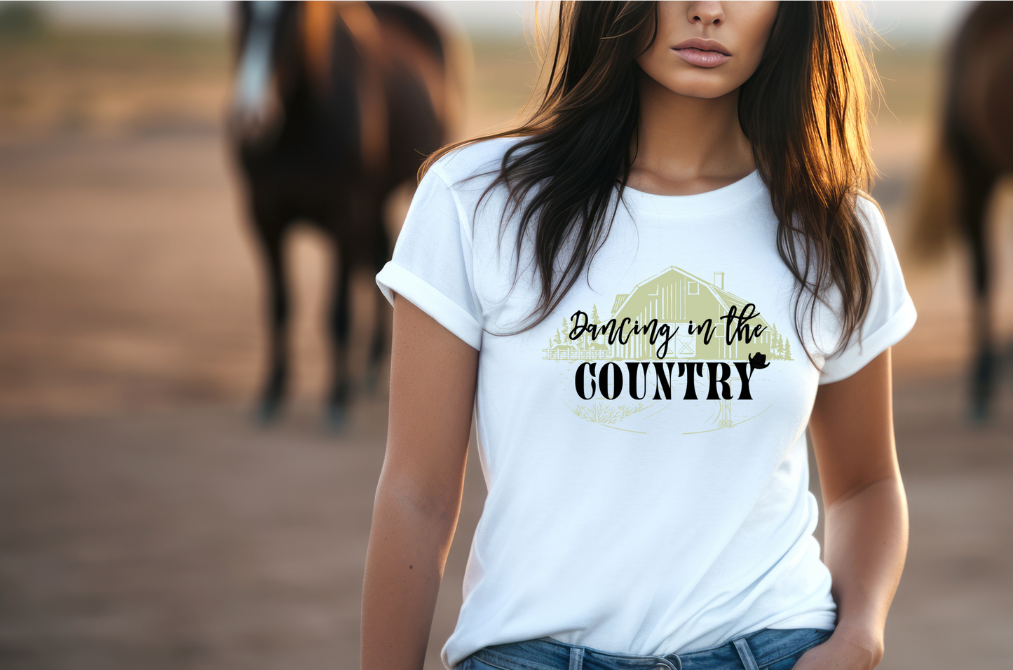 This shirt features a rustic barn set against a serene backdrop of pine trees. The phrase “Dancing in the Country” is styled in a casual, handwritten font and a silhouette of a cowboy hat sit upon the "y"